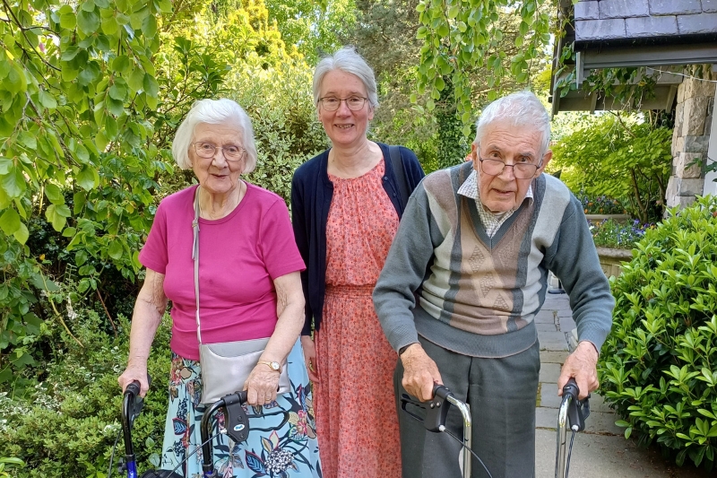Lady standing in the centre between her two elderly parents who are standing with the aid of zimmer frames